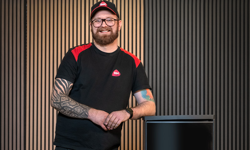 Mann i svart-rødt BM-antrekk med tatoverte armer lener seg mot svart bord foran spilevegg i tre