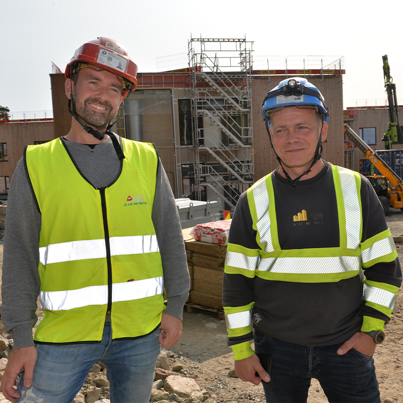 To anleggsarbeidere i gule refleksvester og hjelm foran byggeplass med stillas