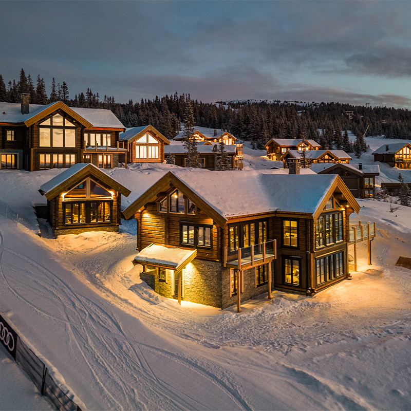 Flere snødekte trehytter med opplyste vinduer i kveldslys ved skogkledd fjellområde.