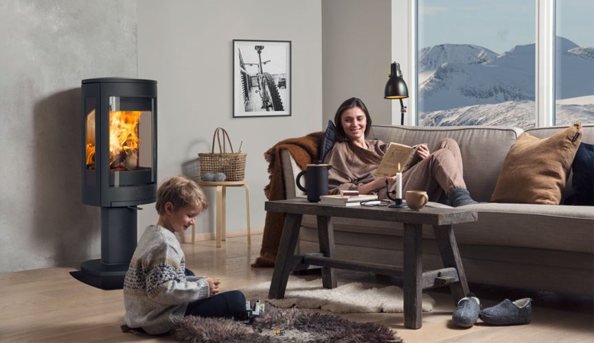 Koselig stue med svart peis, grå sofa, trebord, barn leker på teppet og fjellutsikt.