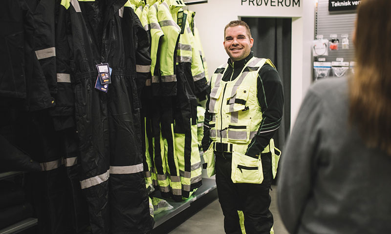 Butikkinteriør med hengende gule refleksdrakter og person i gul sikkerhetsvest ved prøverom.