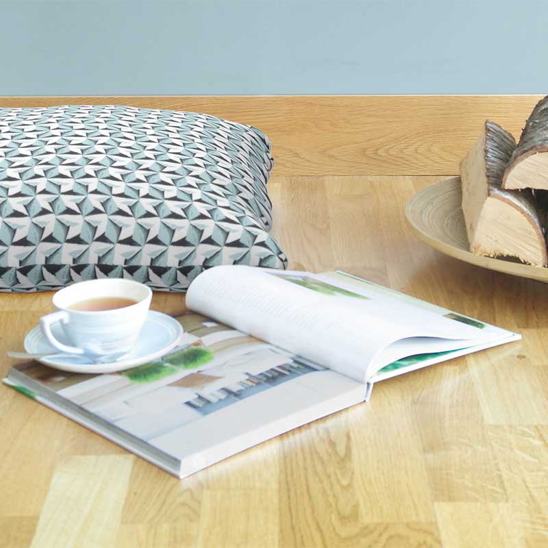 Hvit kopp te på åpen bok på lyst tregulv ved gråhvit geometrisk pute og skål med vedkubber