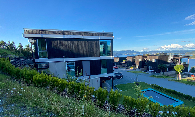 Moderne svart-hvitt bolighus i skråning med grønt gress, blå himmel og vann i bakgrunnen.