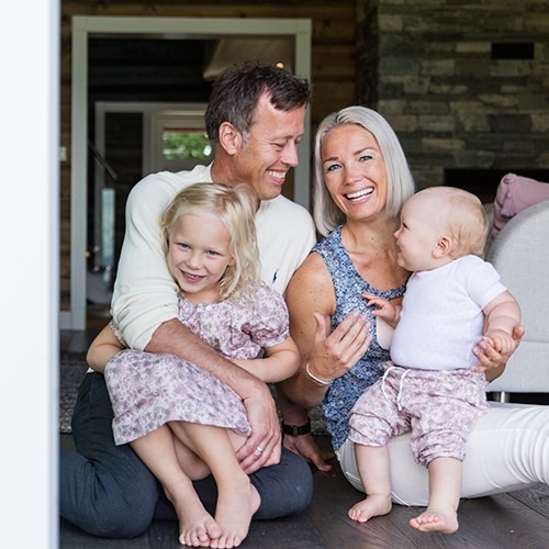 Foreldre med to barn sitter foran grå steinvegg i stue, lyse klær, mørkt tregulv.