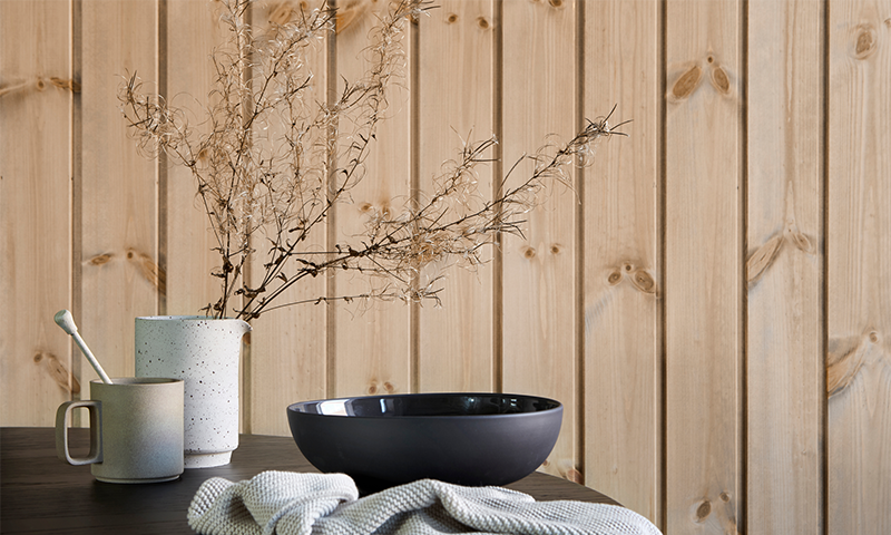 Bord med lys keramikkmugge og -kanne med tørkede strå, svart bolle, strikket klut foran trepanel.