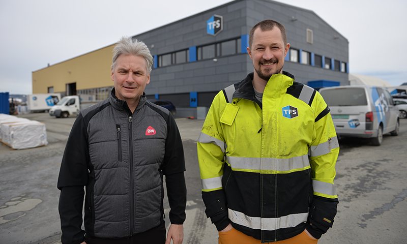 To håndverkere i mørk vest og gul refleksjakke foran grå-gult industribygg med TFS-logo