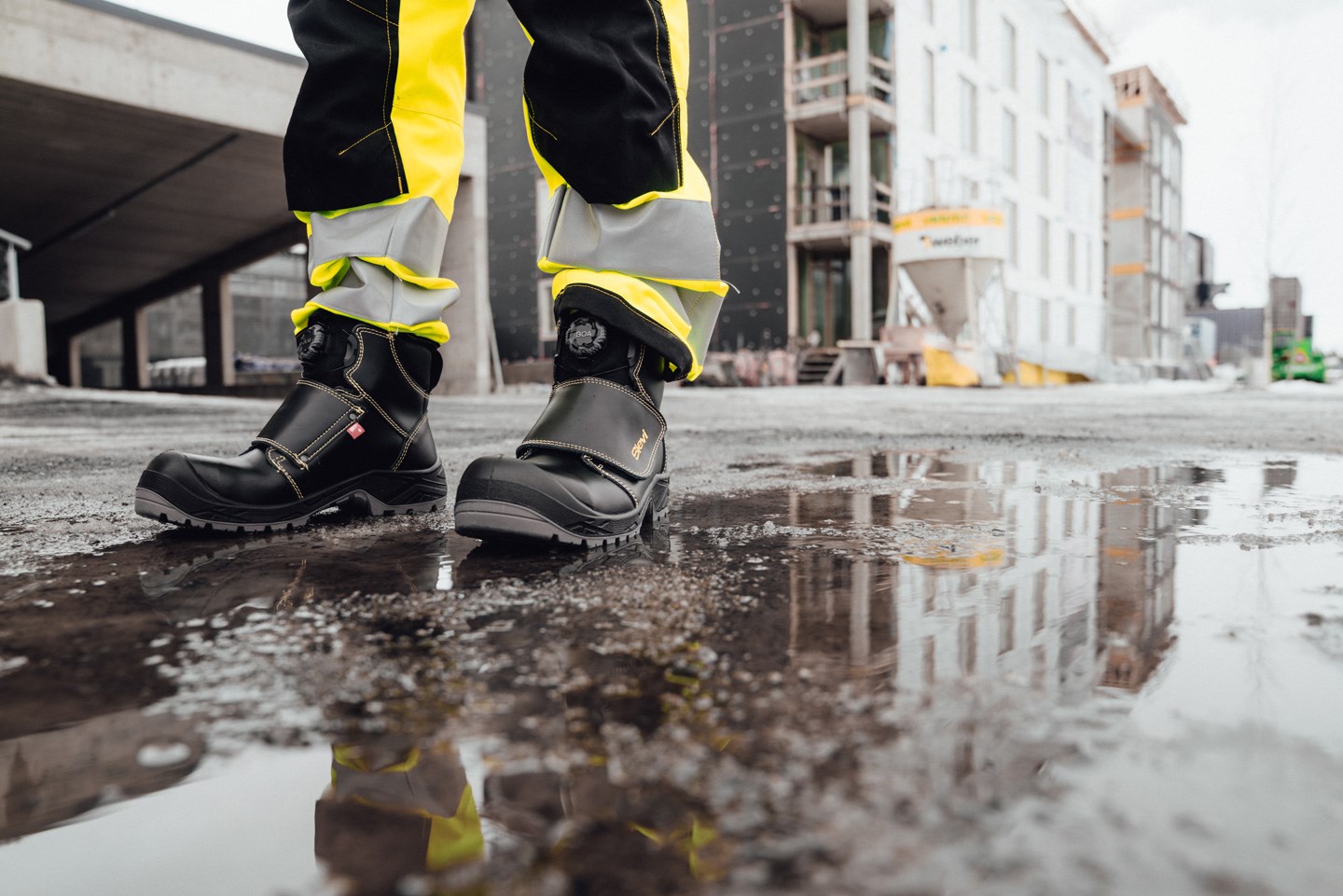 Sorte sikkerhetssko med gule detaljer og refleksbukse i vanndam på byggeplass  