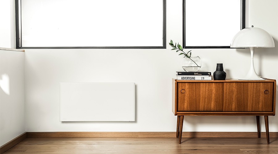 Lyst minimalistisk rom med hvit vegghengt panelovn, teakskjenk med lampe, vase og bøker.