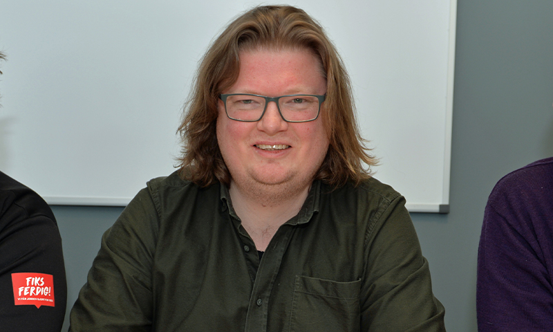 Ung mann med langt blondt hår og grønn skjorte foran hvit tavle i grått møterom.