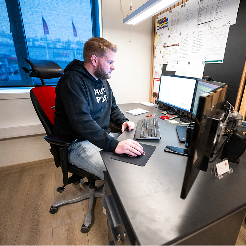 Person i mørk hettegenser ved skrivebord med dobbel skjerm, rød kalkulator og vindu.