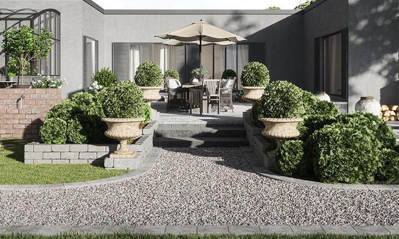 Gårdsrom med grå fasade, grusgang, hevede bed med grønne busker og sitteområde under beige parasoll.