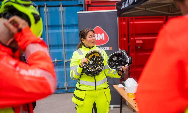 Person i gul refleksjakke holder to hjelmer foran containere og 3M-banner.
