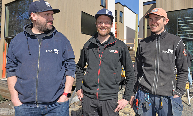 Tre håndverkere i mørke hettejakker og caps foran lyse, moderne hus under oppføring.