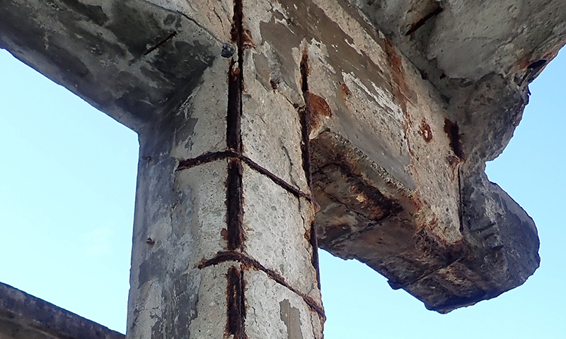 Nærbilde av rustne armeringsjern gjennom sprukken grå betong mot blek blå himmel