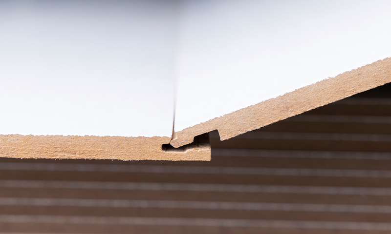 Nærbilde av to hvite malte sponplanker med not- og fjær-skjøt mot brunt underlag.