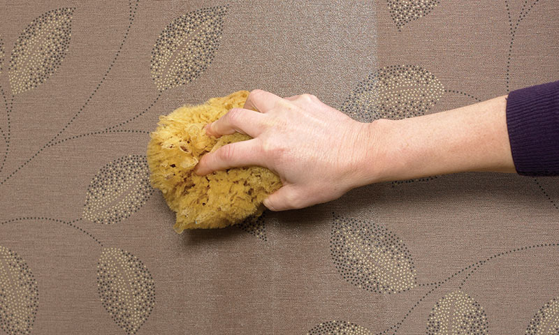 Hånd med gul svamp vasker brun tapet med bladmønster.