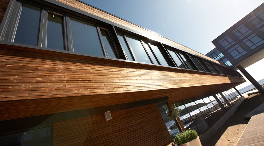 Moderne, langsgående trefasade med store vinduer ved vannkanten under solskinn.