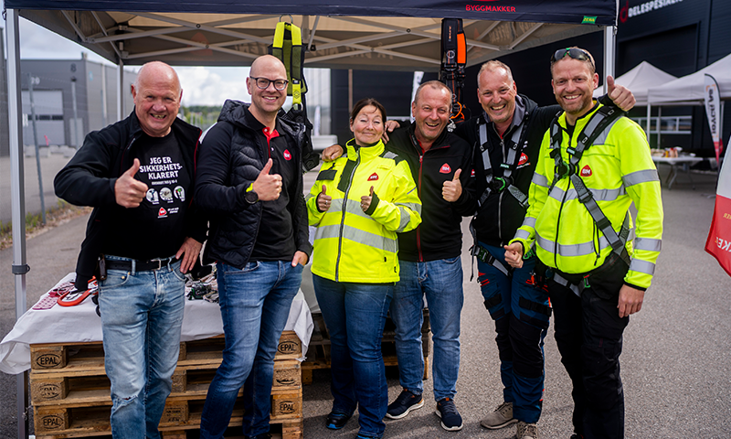 Gruppe på seks i gule refleksjakker under telt med Byggmakker-logo, tommel opp.