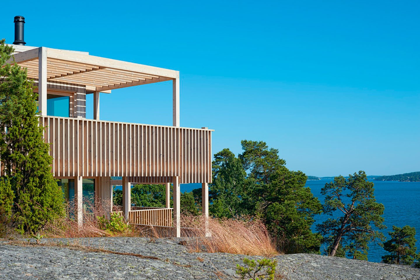 Lys trehytte på berghylle med veranda, furutrær og utsikt mot blått hav