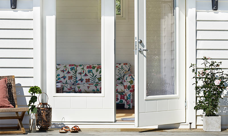 Åpen hvit verandadør til rom med blomstrete sofa, trebenk med rosa pute, lykte, sandaler og rosebusk