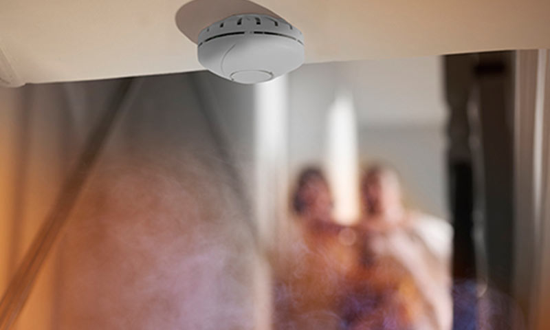 Hvit røykdetektor i taket over seng, varmeoransje lys og stigende røyk