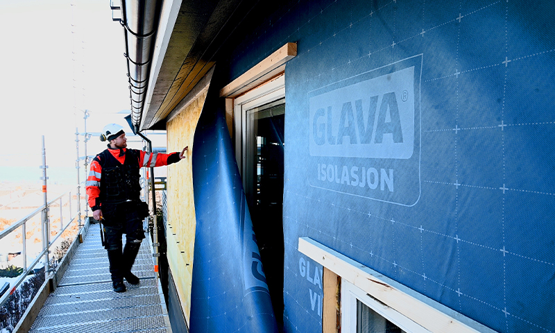 Mann i rød jakke og hvit hjelm sjekker blå Glava-isolasjon på stillas utenfor hus