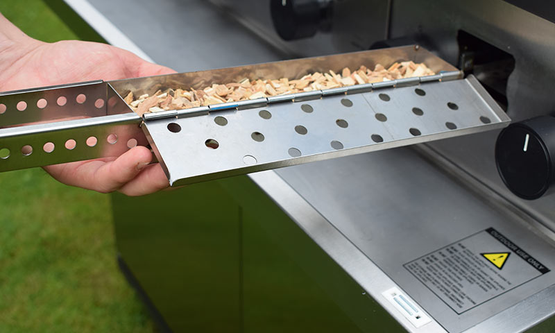 Hånd trekker ut perforert metallbrett med trespon fra rustfri grill på gress.