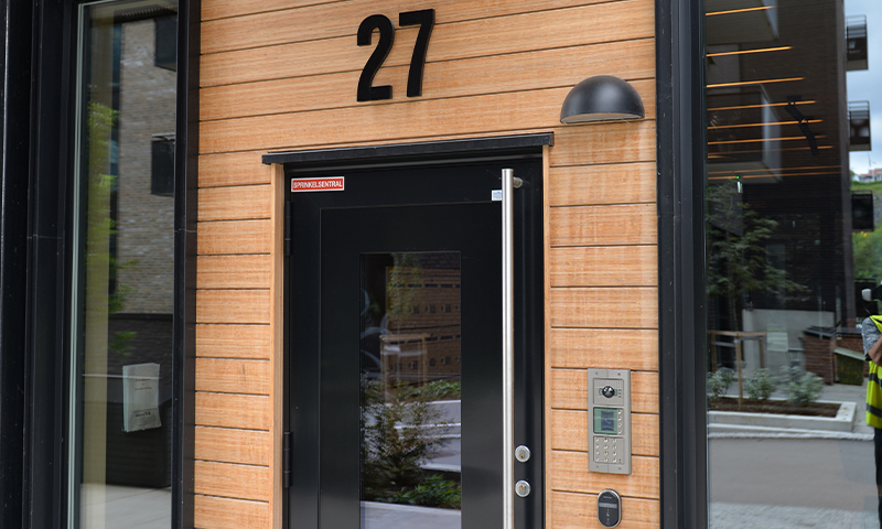 Inngangsparti med svart dør med glass, husnummer 27 på lyst trepanel, ringeklokke og vegglampe.