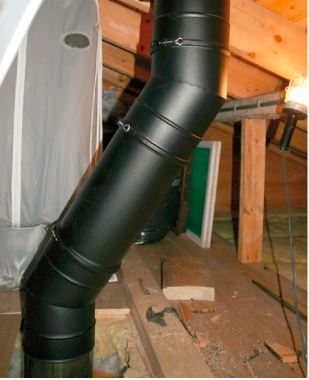 Svart skorsteinsrør i lyst trebjelkelag på loft, omgitt av byggematerialer.