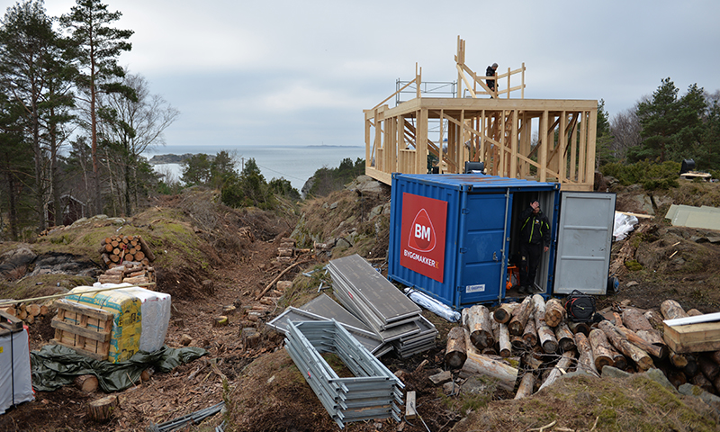 Byggeplass på kystklippe med trekonstruksjon, blå container, tømmerstokker og havutsikt.