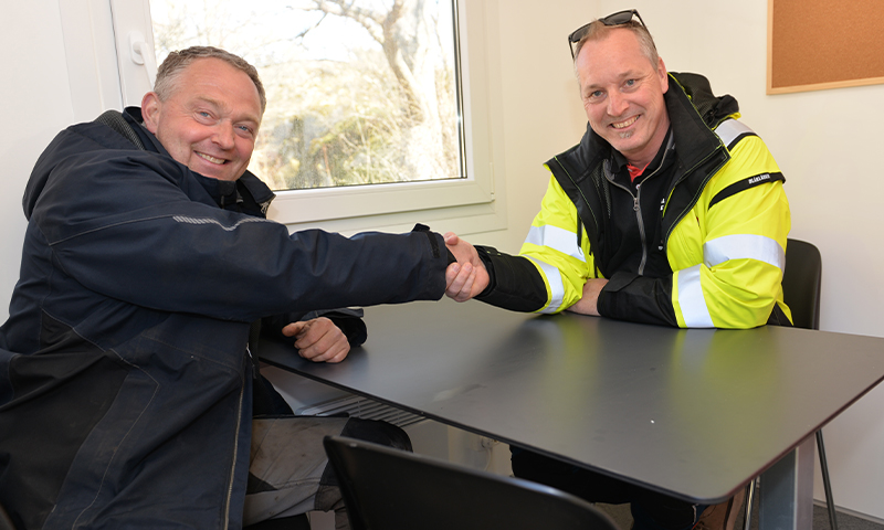 To menn sitter ved et bord inne og hilser med håndtrykk, en med gul refleksjakke og en i mørk jakke