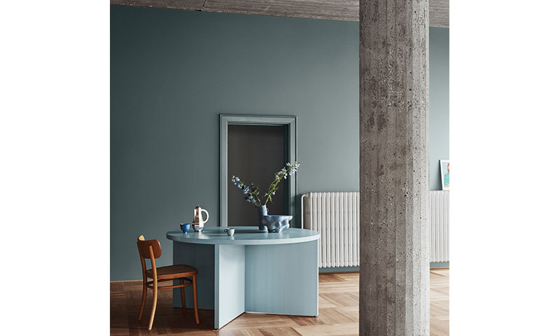 Rundt lyseblått bord med vase og blomster, brun stol, betongsøyle, dempet blå vegg