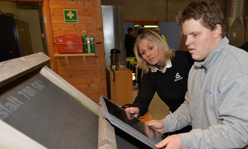 To personer i verksted peker på sort takstein på treramme, oransje vegg med førstehjelpsskilt