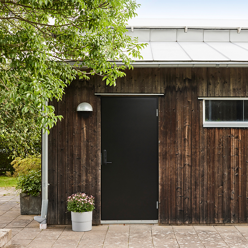 Mørk trebebygning med svart dør, sølvlampe, vindu, potte med rosa blomster og grønt bladverk.