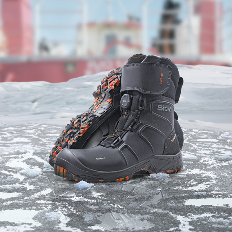 Svart Sievi vinterarbeidsstøvel med oransje mønster på sålen på islagt dekk, båt i bakgrunnen
