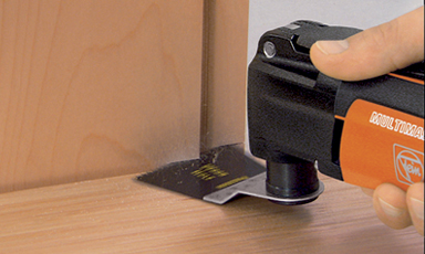 Oransje og svart multiverktøy kutter tre under dørkarm, lys hånd holder det.