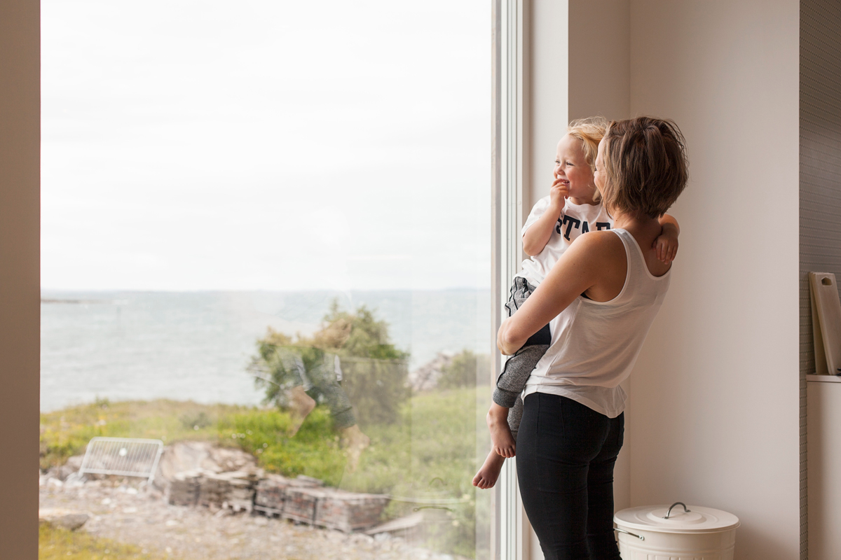 Kvinne i hvit topp holder barn ved stort vindu mot grå sjø og grønne busker.