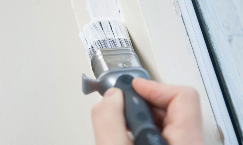 Nærbilde av hånd som maler hvit list ved vindu med grå pensel.