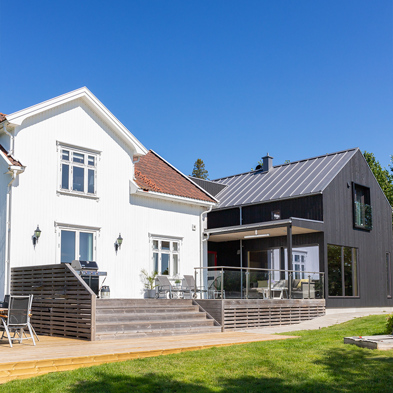Moderne bolig med hvitt tradisjonelt hus og svart tilbygg, stor terrasse med utemøbler under klar blå himmel.