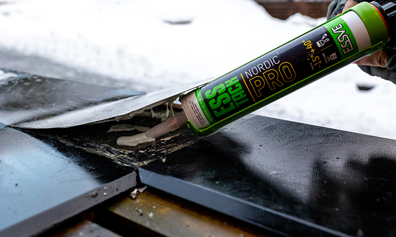 Hånd påfører hvit tetningsmasse fra grønt ESVE Nordic Pro-rør på svart tak med snø.