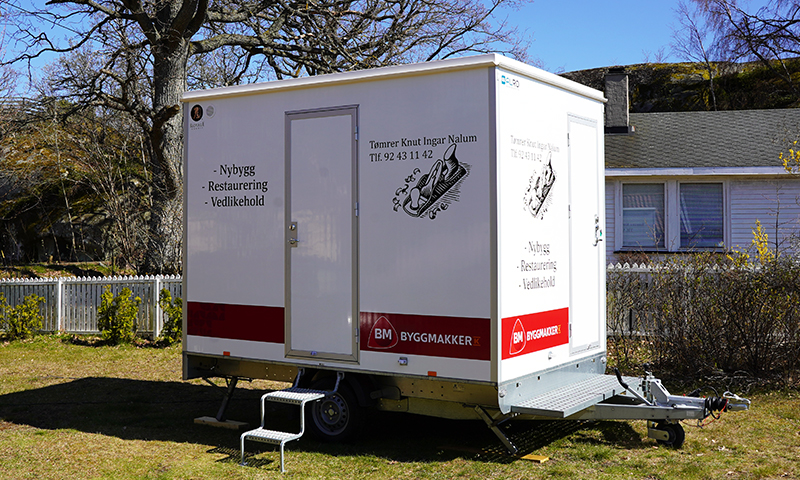 Hvit Byggmakker-vogn med rød stripe på gressplen svart skrift om nybygg, restaurering og vedlikehold.