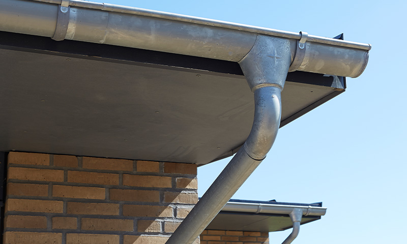 Galvanisert takrenne og nedløpsrør på mursteinsvegg under blå himmel.
