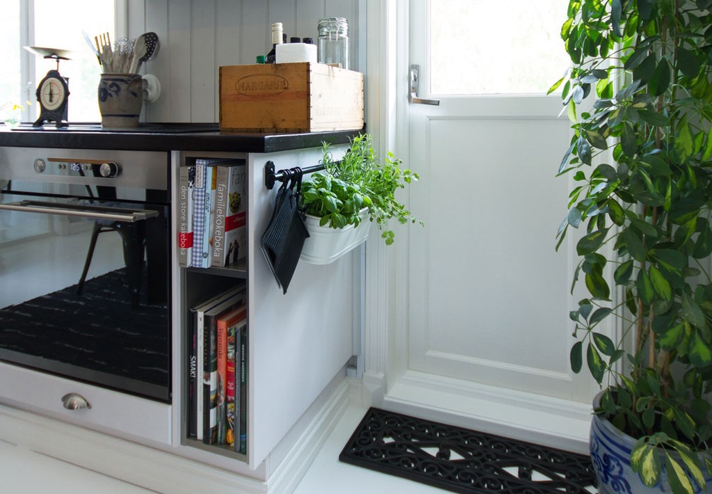 Lyst kjøkken med hvit ovn, trekrate på benk, urter i hvite ampler og grønn plante ved dør