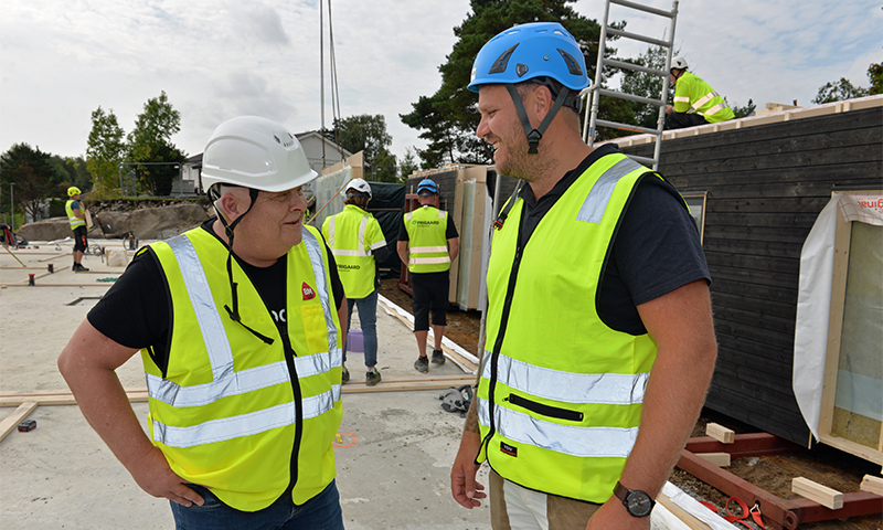 To arbeidere med gule vernevester og hjelmer snakker på byggeplass med stillas og materialer.