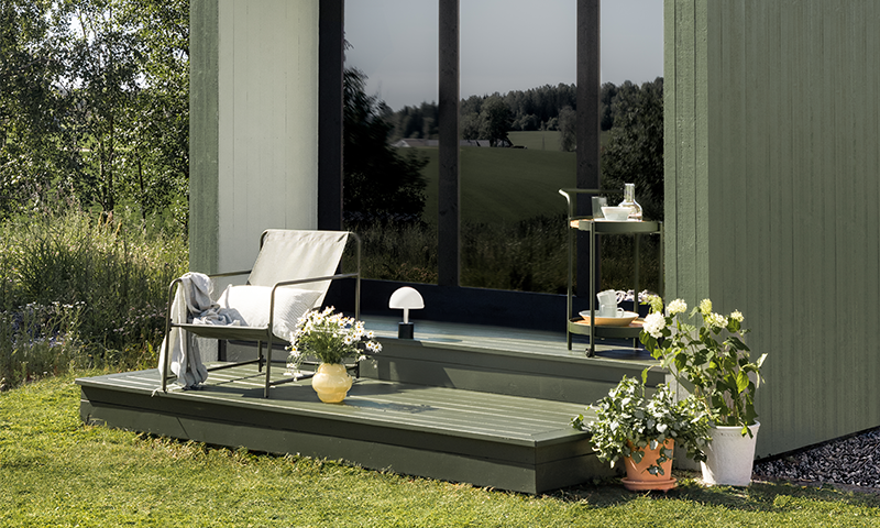 Uteplass ved grønn hytte med lys grå lenestol, gule og hvite blomsterpotter og rullbart bord