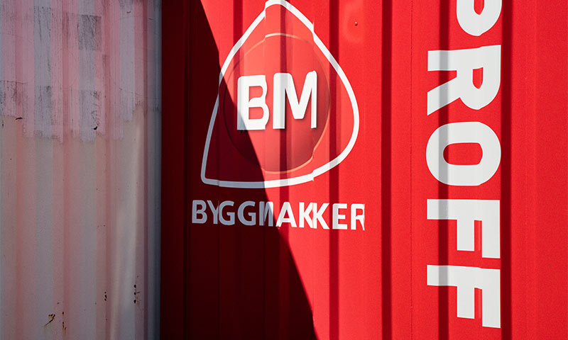 Rød containervegg med hvit BM-logo og teksten Byggmakker Proff, delvis skygge over lyserød panel.