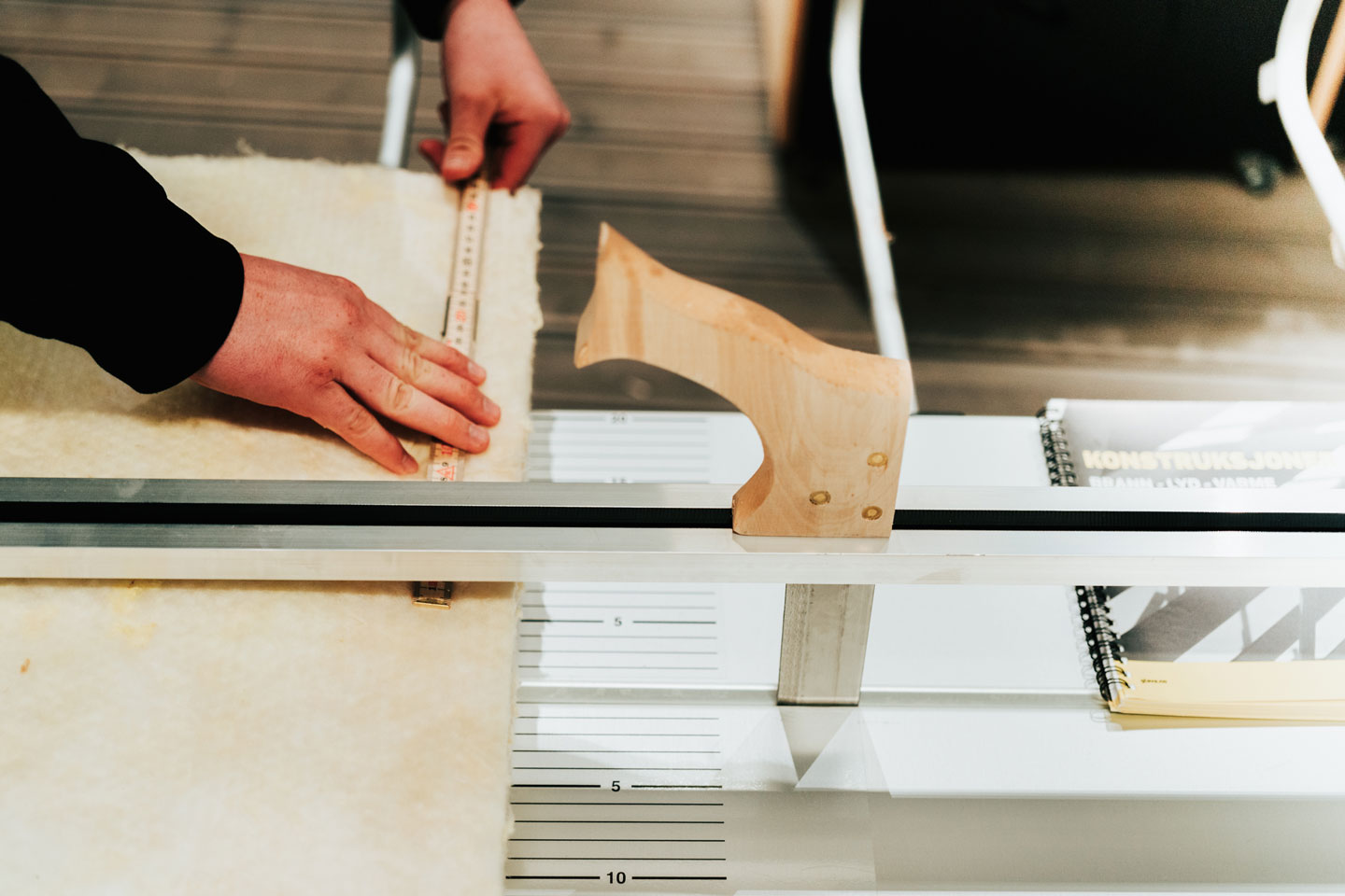 Nærbilde av hender som bruker linjal til å måle og kutte lyst filtstoff på hvitt arbeidsbord.