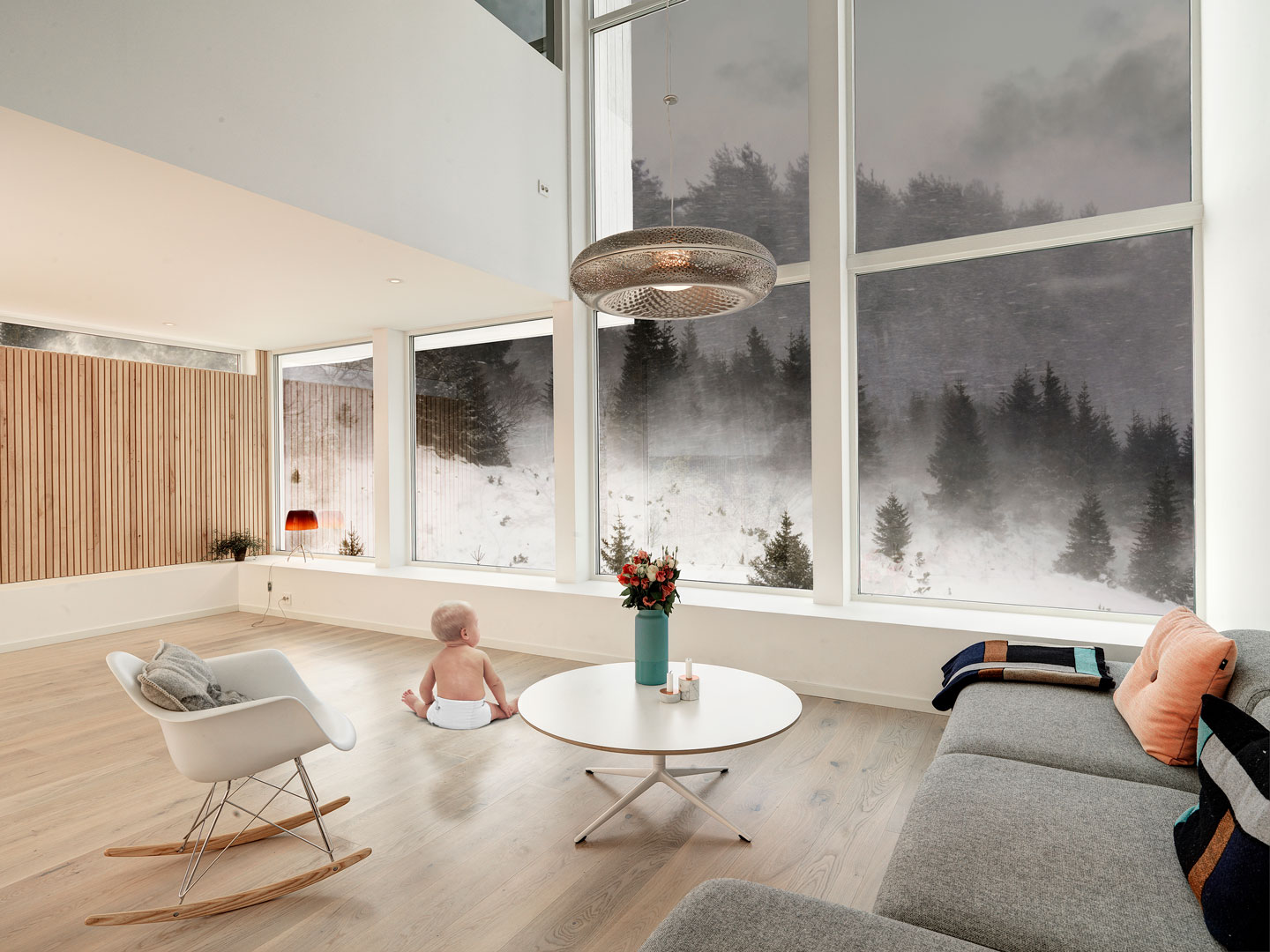 Baby i bleie på lyst tregulv i stue med store vinduer mot snødekt skog og grå sofa