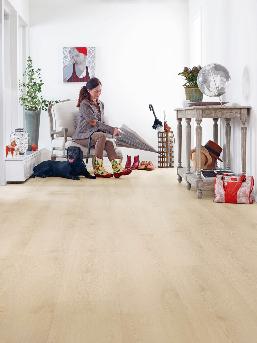 Lyst inngangsparti med dame i mønstret støvler og paraply, hund, konsollbord med hatter og veske.