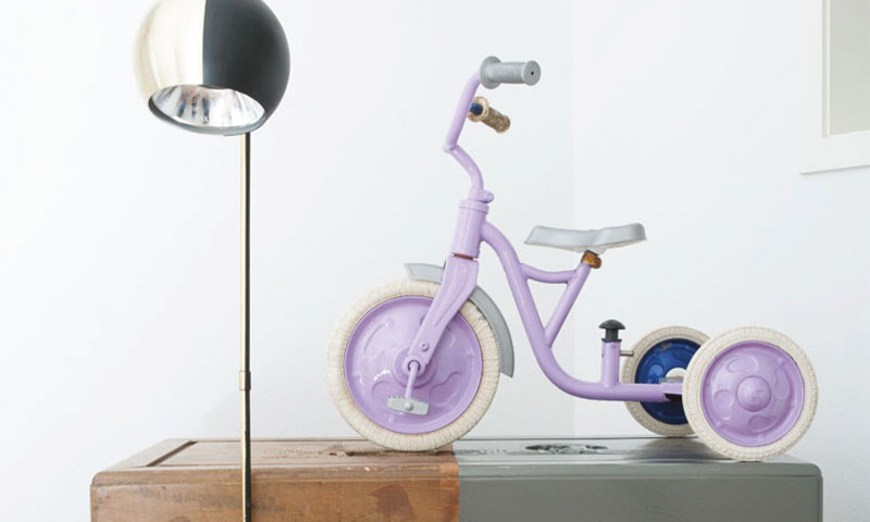 Lilla trehjulssykkel på bord ved sort-hvitt lampe foran hvit vegg.